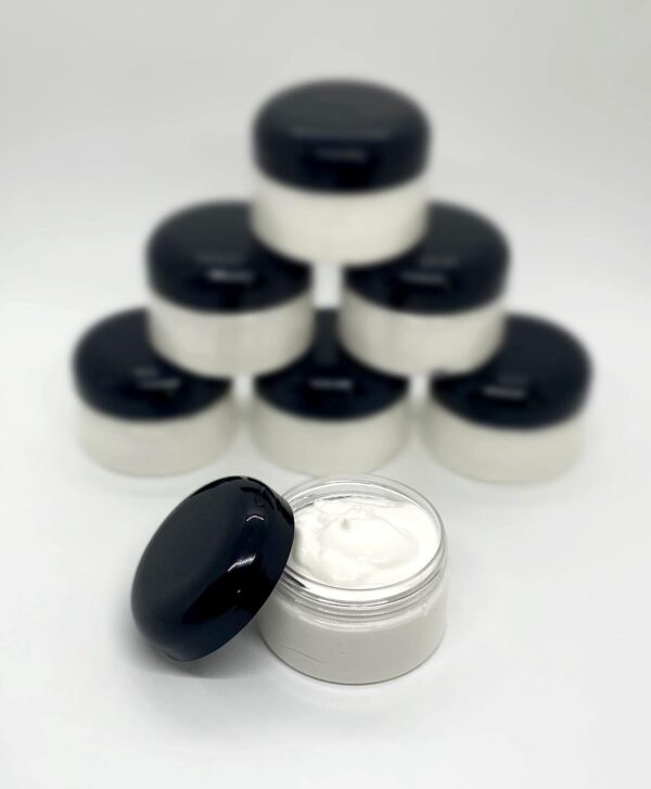 A white container featuring a cream-colored body and a black lid, holding body butter. Stacked jars in the background.